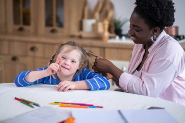 Doing hair. A smiling dark-skinned woman doing hair to the cute girl with down syndrome