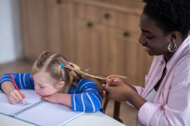 Braids. A positive happy woman making braids to a cute girl sitting next to her