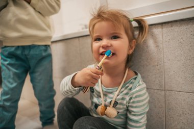 Cute female child sitting in the bathroom and brushing her teeth in the presence of her sibling