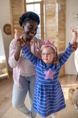 Happy moments. Smiling dark-skinned lady having good time with a cute girl in blue dress