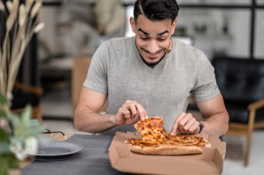 Favorite food. Joyful young bearded man with looking at fresh pizza taking slice sitting at table in apartment