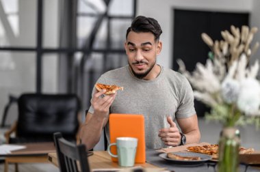 Good time. Chewing smiling young man holding piece of pizza looking curiously surprised at tablet sitting at table