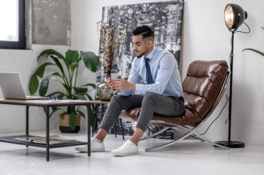 Waiting. Successful attractive young man in shirt with tie sitting in armchair looking at smartphone in room during daytime