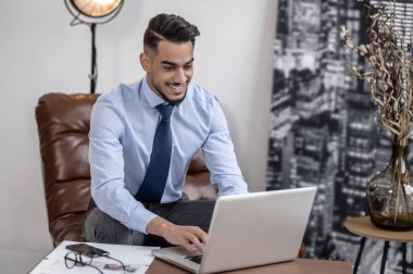 Inspiration. Business young bearded man in tie smiling typing on laptop sitting in leather armchair indoors