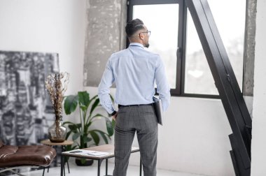 Confidence. Business man in white shirt with laptop standing with back to camera looking out window in room during day