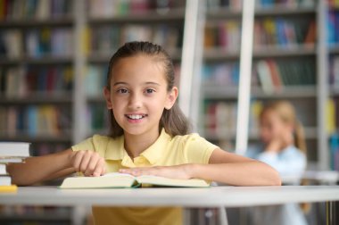 Portrait of a smiling cheerful cute adolescent girl sitting with an open book at the library table