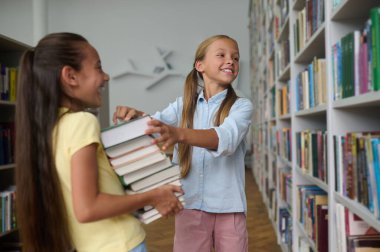 Smiling cute teenage girl helping her cheerful classmate to select textbooks in the reading room
