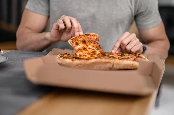 Eating pizza. Hands of man sitting at table at home taking slice of delicious pizza without face