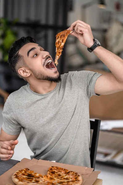 Appetizing pizza. Young bearded hungry man holding piece of pizza over open mouth while sitting at table at home