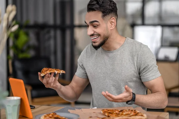 Time for yourself. Young attractive man with piece of pizza gesturing smiling looking at tablet screen sitting at table at home