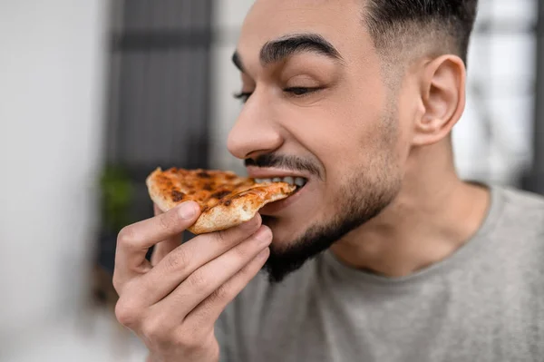 Tasty food. Face of young bearded man sideways to camera biting off delicious fresh pizza indoors