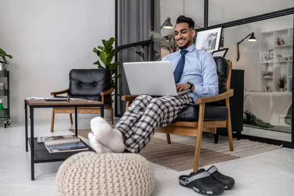 Work remotely. Young bearded smiling man in tie working on laptop sitting at home in comfortable leather chair