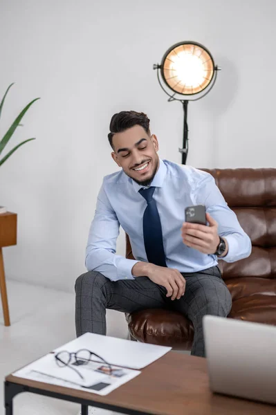 Lucky day. Confident young business man smiling looking at smartphone sitting in modern office