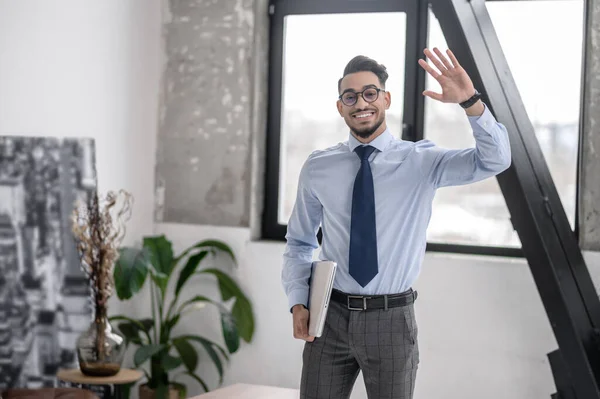 Greetings. Smiling young bearded man in glasses with laptop raising hand in greeting walking towards camera in office