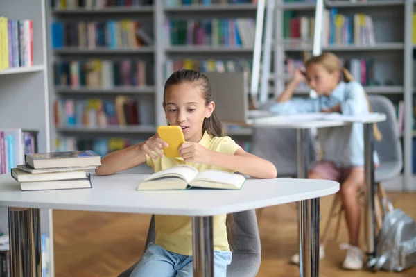 Focused Dark Haired Teenage Girl Sitting Library Table Looking Smartphone — Zdjęcie stockowe