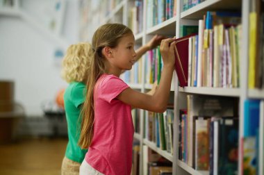 Focused cute long-haired teenage girl and a blond boy standing before the bookshelves the school library