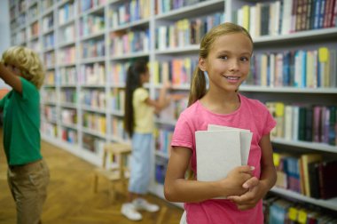 Joyous pretty teenage girl with a pile of book in her hands and her friends standing among the bookshelves