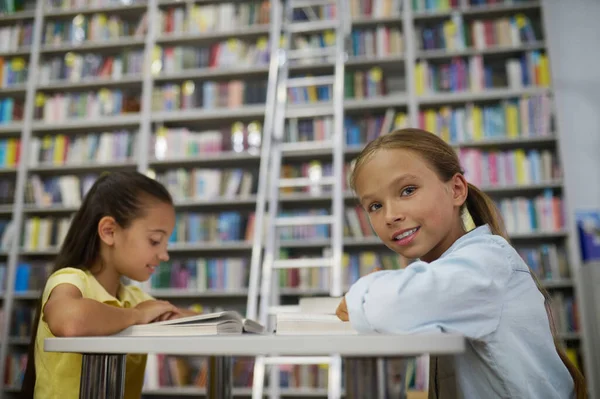 Smiling Pleased Schoolgirl Her Focused Friend Sitting Table School Library — Zdjęcie stockowe