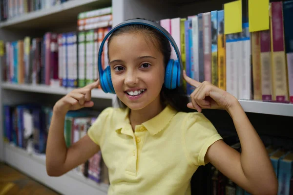 Waist-up portrait of a pleased young lady in the wireless headphones standing at the shelving unit in the library