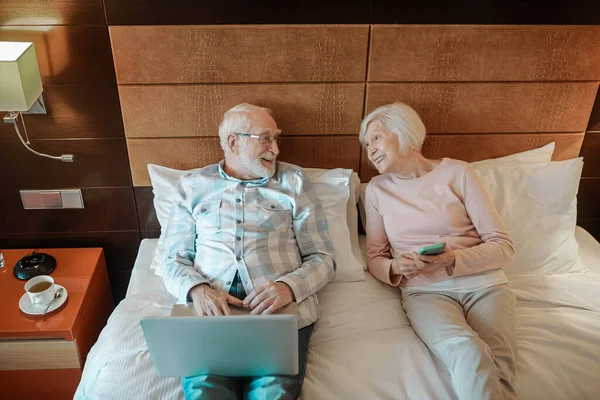 Resting in a hotel. Senior couple lying on bed in a hotel room and watching something online