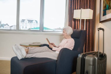 A senior woman in pink blouse with a tablet sitting in an armchair
