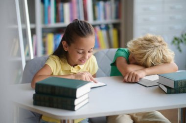 Smiling focused schoolgirl seated at the table reading while a schoolboy dozing on an open textbook