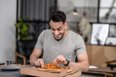 Snack. Joyful young adult man touching pizza in box sitting at table at home