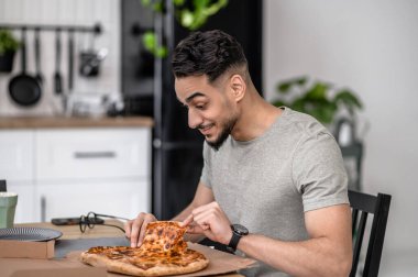 Eating at home. Happy young bearded man touching pizza while sitting sideways to camera in apartment