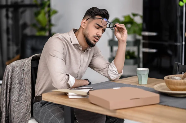 Short break. Attractive young bearded man with closed eyes taking off glasses sitting at table at home