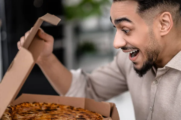 Hungry man. Young bearded man with open mouth sideways to camera looking enthusiastically at pizza in open box indoors