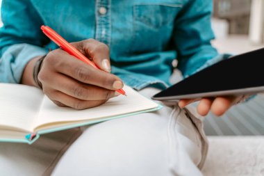 Notes. Hands of dark-skinned man sitting outdoors writing with pen and holding tablet without face