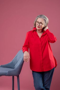 In a studio. Mature woman in red shirt and eyeglasses standing near the chair