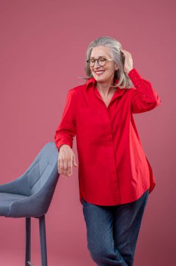 In a studio. Mature woman in red shirt and eyeglasses standing near the chair