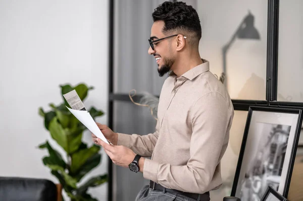 Good news. Smiling attractive man in glasses reading documents standing sideways to camera in apartment