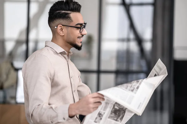 Interest. Smiling young bearded man in glasses standing sideways to camera reading newspaper with interest indoors
