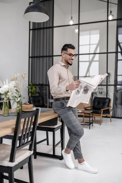 Latest news. Young stylish man in glasses standing leaning on table reading newspaper in cozy room at home