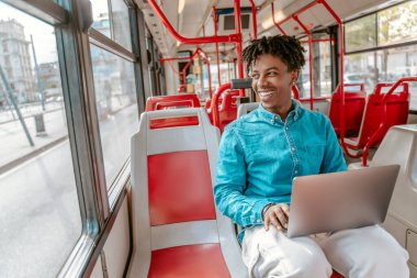 Lucky day. Happy african guy sitting with open laptop in city bus smiling looking out window on sunny day