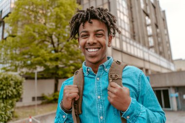 All excellent. Smiling african guy with backpack looking at camera confidently standing on city street on fine day