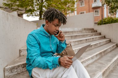 Always in touch. Focused serious dark-skinned guy talking on smartphone working on laptop sitting on steps outdoors