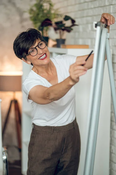 Working on project. Mature dark-haired woman standing near the flipchart and working on project details