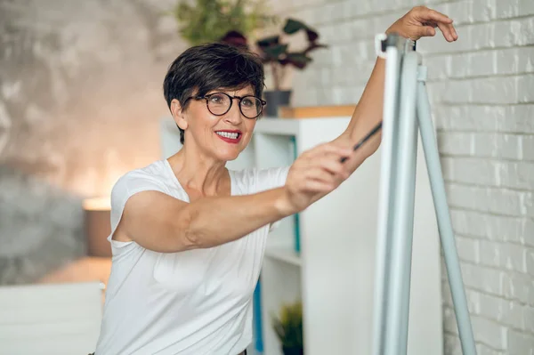 Working on project. Mature dark-haired woman standing near the flipchart and working on project details