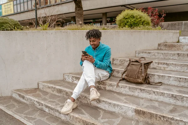 Time for yourself. Smiling african man looking interested in smartphone sitting near backpack on steps outdoors on fine day