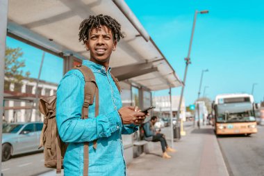 Waiting. Young african man with smartphone and backpack standing at bus stop looking ahead on city street on sunny day