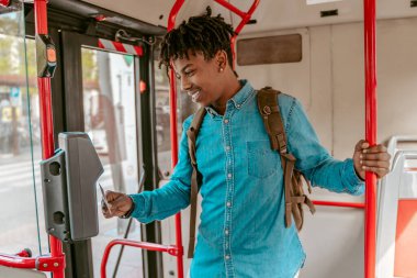 Travel payment. Dark-skinned friendly guy with backpack standing holding handrail bringing card to reader in city bus