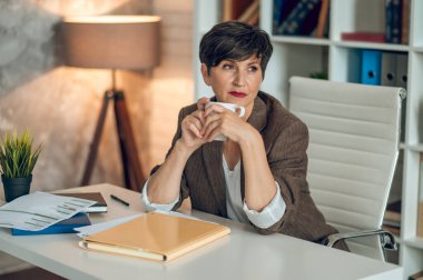 Morning in the office. Good-looking dark-haired woman sitting at the table with a coffee mug in hands