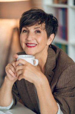 Morning in the office. Good-looking dark-haired woman sitting at the table with a coffee mug in hands