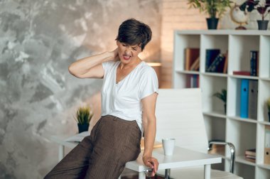 Neck pain. Dark-haired woman in the office feeling bad and suffering from neck pain