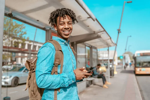 Lucky day. African guy standing sideways smiling at camera holding smartphone at bus stop on city street on sunny day