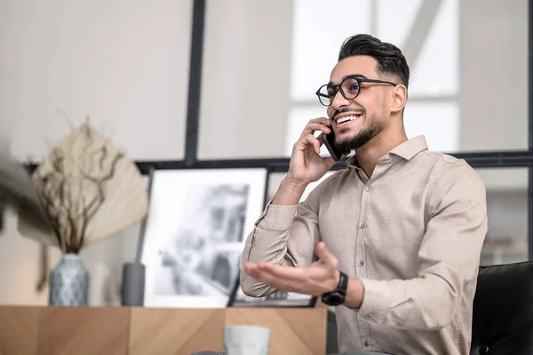 Phone call. Smiling young bearded elegant man gesturing talking on smartphone while sitting indoors