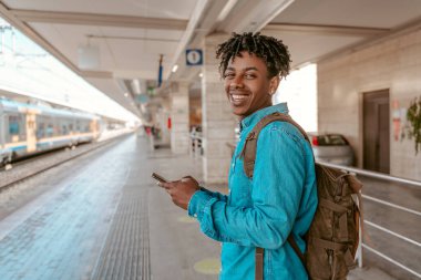 Lucky day. African guy standing sideways smiling at camera holding smartphone on station platform and train on railway